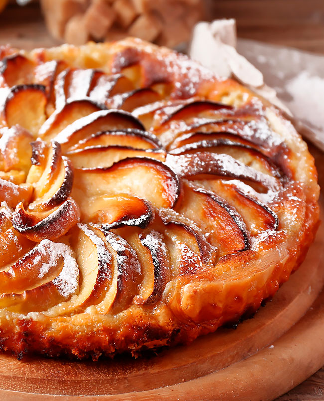 Tarte de Maçã da Avó Tarte de Maçã da AvóA tarte de maçã à moda antiga pertence precisamente a essa categoria. Tem o perfume doce da maçã assada, o calor reconfortante da canela e aquela massa dourada que se desfaz delicadamente a cada garfada. É uma sobremesa simples, humilde e genuína, mas capaz de transformar uma tarde banal num momento especial. Quem teve a sorte de crescer com uma avó que gostava de cozinhar talvez reconheça imediatamente este aroma. A tarte acabada de sair do forno, a cozinha aquecida, a mesa preparada e uma chávena de café ou chá à espera. São memórias que não precisam de fotografias para permanecerem vivas. E embora nenhuma receita consiga reproduzir exatamente o sabor das mãos de uma avó, esta tarte de maçã à moda antiga aproxima-se bastante desse conforto. Tarte de maçã à moda antiga: um clássico que nunca passa de moda A grande beleza desta receita está na simplicidade. Não são necessários ingredientes sofisticados nem técnicas complicadas. O segredo está em respeitar cada etapa, escolher bons ingredientes e, sobretudo, não ter pressa. A combinação entre maçã, canela, açúcar, leite, ovos e massa quebrada resulta numa sobremesa cremosa por dentro, dourada por fora e irresistivelmente aromática. É também uma excelente forma de aproveitar maçãs que estejam maduras, desde que ainda se encontrem em boas condições. A tarte pode ser servida morna, à temperatura ambiente ou fria. Contudo, há algo de especial em provar a primeira fatia ainda ligeiramente quente, quando o aroma da maçã e da canela permanece no ar. O segredo começa na escolha das maçãs Nem todas as maçãs produzem exatamente o mesmo resultado. Para uma tarte equilibrada, podem ser utilizadas maçãs com alguma acidez e boa firmeza, pois ajudam a contrariar a doçura do açúcar e mantêm uma textura agradável durante a cozedura. As maçãs devem estar maduras, mas não demasiado moles ou deterioradas. Uma fruta de qualidade é essencial para qualquer sobremesa. Depois de descascar e cortar as maçãs, existe ainda um pequeno truque que faz diferença no resultado final: regar as fatias com algumas gotas de sumo de limão. Além de acrescentar uma ligeira nota de frescura, o limão ajuda a evitar que a maçã oxide e fique escura antes de chegar ao forno. Massa quebrada ou massa folhada? A receita tradicional apresentada utiliza massa quebrada, que proporciona uma base delicada e ligeiramente estaladiça. É uma escolha particularmente feliz para esta tarte porque permite que o recheio de maçã se destaque. No entanto, quem preferir uma textura mais leve e folhada pode optar por massa folhada. O resultado será diferente, mas igualmente apetecível. Seja qual for a escolha, é importante picar o fundo da massa com um garfo antes de adicionar o recheio. Este pequeno gesto ajuda a evitar que a massa cresça excessivamente durante a cozedura. O forno é parte do segredo Um dos erros mais frequentes na preparação de sobremesas é colocar a tarte num forno ainda frio. Para conseguir uma massa bem cozida e uma superfície dourada, o forno deve ser pré-aquecido a 200 ºC. O choque térmico inicial contribui para que a massa comece a cozinhar imediatamente e ajuda a criar a textura desejada. À medida que a tarte assa, a cozinha começa a encher-se daquele perfume inconfundível de maçã e canela. É precisamente nesse momento que esta receita deixa de ser apenas uma sobremesa e começa a transformar-se numa memória. Como conseguir uma tarte bonita e dourada A apresentação também conta. As fatias de maçã devem ser cortadas finamente e dispostas cuidadosamente sobre a massa. Uma forma clássica consiste em colocá-las em círculos, começando pelo rebordo da tarteira e avançando progressivamente até ao centro. Além de proporcionar uma apresentação mais elegante, esta disposição permite distribuir a maçã de forma relativamente uniforme. No final, o creme é vertido sobre as maçãs, envolvendo-as durante a cozedura. O resultado deverá ser uma tarte dourada, perfumada e com um interior macio. Tarte de maçã à moda antiga, como a avó fazia Ingredientes 4 maçãs 3 ovos 1 embalagem de massa quebrada 50 g de farinha 150 g de açúcar, de preferência amarelo 1 pitada de canela 1 chávena de leite Algumas gotas de sumo de limão Modo de preparação Comece por reunir todos os ingredientes e utensílios necessários. Esta organização inicial torna a preparação muito mais simples. Pré-aqueça o forno a 200 ºC. Forre uma tarteira com a massa quebrada, mantendo o papel vegetal que acompanha a massa, se existir. Pique o fundo e as laterais da massa com um garfo. Este passo ajuda a impedir que a base cresça demasiado durante a cozedura. Lave cuidadosamente as maçãs. Corte-as em quartos, retire a casca e elimine os caroços. Corte as maçãs em meias-luas finas. Regue ligeiramente as fatias com algumas gotas de sumo de limão para ajudar a preservar a sua cor. Disponha as fatias de maçã sobre a massa, formando filas circulares desde o exterior até ao centro. Num recipiente alto, coloque os ovos, o leite, a farinha, o açúcar e uma pitada de canela. Triture tudo com a varinha mágica até obter um creme homogéneo. Verta cuidadosamente o creme sobre as maçãs, procurando distribuí-lo de forma uniforme. Leve a tarte ao forno durante aproximadamente 35 minutos. Vigie a cozedura. O tempo pode variar consoante o forno e a altura da tarte. Para verificar se está pronta, faça o teste do palito numa zona de creme. Se sair praticamente seco, a tarte estará cozida. Nos minutos finais, deixe a superfície ganhar uma bonita tonalidade dourada, tendo atenção para não deixar queimar. Retire do forno e deixe repousar alguns minutos antes de cortar. Sirva morna ou à temperatura ambiente. O resultado é uma tarte de maçã cremosa, aromática e delicadamente dourada, com aquele sabor caseiro que parece transportar-nos diretamente para a cozinha da avó. Como deixar a tarte ainda mais saborosa Há pequenos pormenores capazes de elevar esta receita. Canela sem exageros A canela deve perfumar a tarte, não esconder o sabor da maçã. Uma pequena quantidade é suficiente para criar aquele aroma característico das sobremesas tradicionais. Açúcar amarelo O açúcar amarelo acrescenta uma nota mais profunda e caramelizada ao conjunto. É particularmente interessante quando combinado com maçãs ligeiramente ácidas. Maçã bem distribuída Não amontoe demasiado as fatias. Uma distribuição equilibrada permite que o calor chegue melhor a toda a fruta e proporciona uma textura mais uniforme. Não corte imediatamente Depois de sair do forno, a tarte ainda está muito quente e o recheio encontra-se bastante delicado. Deixar repousar alguns minutos ajuda a estabilizar a textura e facilita o corte. O toque final: uma tarte com sabor a memória Há receitas que sobrevivem porque são perfeitas. Outras sobrevivem porque significam alguma coisa para alguém. A tarte de maçã à moda antiga pertence às duas categorias. É económica, simples e relativamente fácil de preparar, mas oferece uma combinação de sabores que atravessou gerações. A maçã cozinhada lentamente, o perfume da canela, a massa dourada e o creme delicado fazem desta sobremesa um verdadeiro clássico da cozinha caseira. Pode ser apresentada num almoço de família, num lanche de domingo ou numa ocasião especial. E pode também ser feita simplesmente porque apetece recuperar aquele sabor que, durante alguns minutos, parece fazer o relógio andar para trás. Porque algumas sobremesas não se comem apenas com uma colher. Comem-se com a memória. E uma boa tarte de maçã, feita à moda antiga, tem precisamente esse poder: transformar ingredientes simples numa pequena viagem à infância.